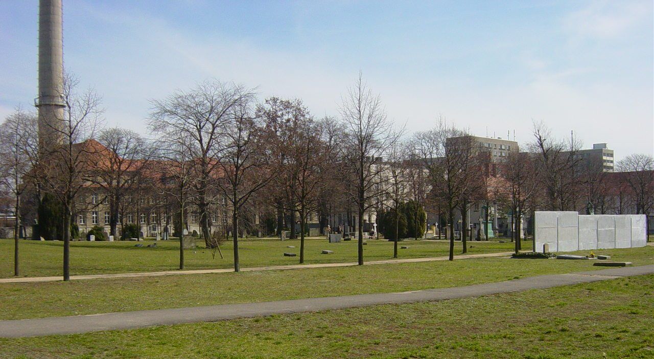 Weite Rasenfläche mit vereinzelten Grabsteinen, Bäumen und einem erhaltenen Abschnitt der Berliner Mauer